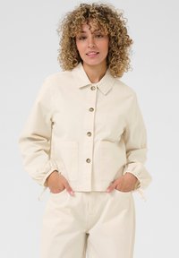 Beige cropped jasje met een kraag, knoopsluiting aan de voorkant en elastische manchetten. Beschikt over twee voorzakken en een gladde stoffen textuur.