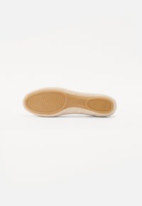 Semelle d'une chaussure, avec une base en caoutchouc texturé de couleur beige. Un matériau doux semblable à du daim recouvre le contour supérieur sans lacets visibles.