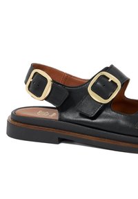 Black leather sandal with two large gold-tone buckles, flat sole, and brown accent on the insole, featuring a textured finish.