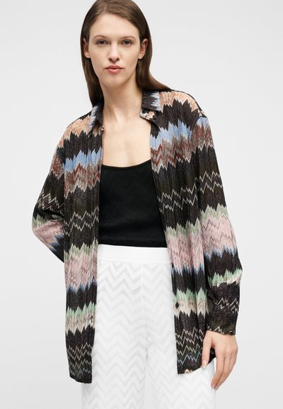 Νεαρή γυναίκα φοράει πολύχρωμο πουκάμισο με ζιγκ ζαγκ μοτίβο και μακριά μανίκια πάνω από μαύρο τοπ και λευκή υφασμάτινη φούστα, στέκεται μπροστά σε απλό φόντο.