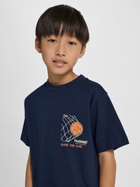 Młody chłopiec z ciemnymi włosami ubrany w granatowy t-shirt z pomarańczową piłką złapaną w siatkę oraz napisem „hummel SHARE THE GAME.”