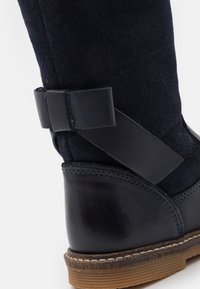 Bota de ante azul marino con punta redondeada, detalles de cuero, suela de goma con costuras, y una correa decorativa de cuero en el talón.