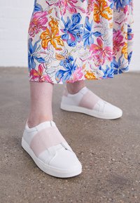 Bílé slip-on boty mají růžový elastický popruh přes střed chodidla s vyraženým logem značky. Hladký materiál, zaoblená špička a plochá podrážka.