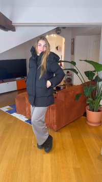 Schwarzer Steppmantel mit Kapuze, zwei Fronttaschen und einer lockeren Passform. Kombiniert mit hellgrauen Hosen und schwarzem Schuhwerk, inszeniert in einem gemütlichen Innenraum.