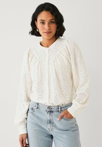 Blusa bianca con tessuto testurizzato a motivo a diamante, chiusura con bottoni e maniche a sbuffo. Abbinata a jeans azzurri a vita alta.
