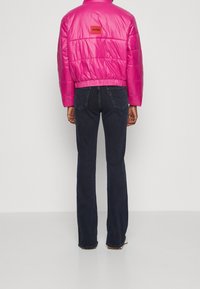 Chaqueta acolchada fucsia con diseño corto, dobladillo elástico y parche con logo en la espalda, combinada con vaqueros acampanados azul oscuro. Textura lisa y brillante.