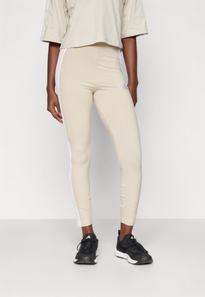 Beige leggings laget av elastisk stoff, med hvite sidestriper og et Adidas-logo på venstre lår. Brukt med svarte treningssko.