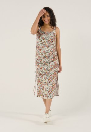 Pregnant woman smiling, wearing a floral sleeveless midi dress and white sneakers, touching her hair while standing on a plain background.