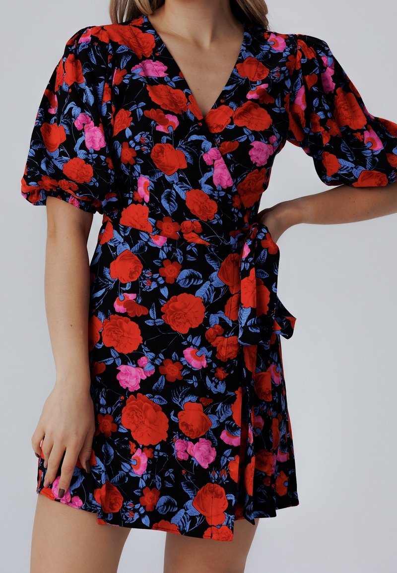 Femme portant une robe portefeuille noire avec des motifs floraux rouges et roses ainsi que des manches courtes bouffantes, posant avec une main sur la hanche.