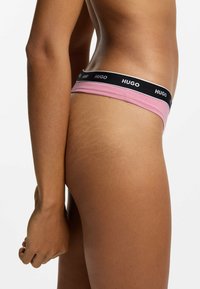Lyserød kvindestring med sort elastikbånd, der har hvid "HUGO"-brandning. Glat stoftekstur og design med minimal dækning.