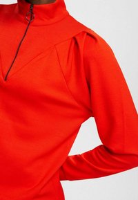 Czerwony sweter z wysokim kołnierzem i zamkiem, z bufkami na ramionach oraz teksturowanym, gładkim materiałem.
