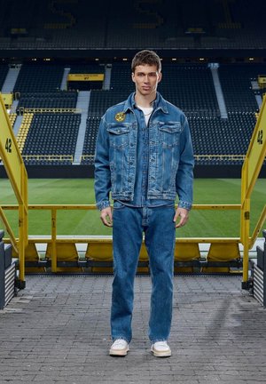 Junger Mann in blauer Jeansjacke und Jeans steht auf dem Stadionweg mit grünem Feld und leeren gelb-schwarzen Sitzen hinter ihm.