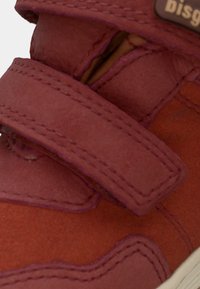 Zbliżenie na czerwoną zamszową sneaker z szerokim paskiem na rzep i widocznymi detalami szycia.