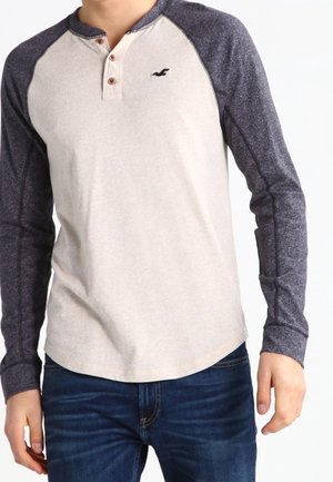 Homme portant un t-shirt henley à manches longues beige et gris foncé avec trois boutons et un jean bleu, debout sur fond blanc.