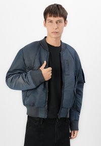 Giacca bomber blu realizzata in materiale lucido e morbido con bordi a costine, dotata di chiusura frontale a zip e una tasca a toppa sul braccio.