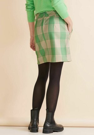 Femme portant un pull vert, une jupe à carreaux beige et verte, des collants noirs et des bottines noires, debout avec une main dans la poche.