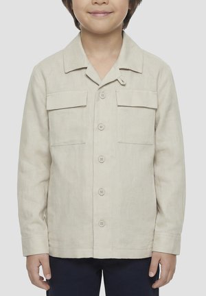 Dijete koje nosi bež jaknu s gumbima i dva džepa s preklopom na prsima te tamne hlače, stoji na jednobojnoj pozadini.