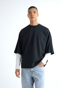 Čierny oversized pulóver s krátkym rukávom, vrstvený cez bielu košeľu s dlhým rukávom, v kombinácii s svetlomodrými džínsami.