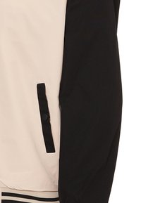 Giacca leggera con corpo beige e maniche nere, dotata di una tasca laterale visibile e orlo a coste a righe. Tessuto morbido, design minimalista.