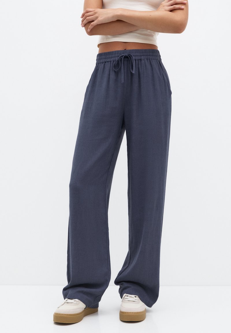 Femme portant un pantalon ample bleu foncé avec cordon et des baskets blanches, debout les bras croisés devant un fond blanc uni.