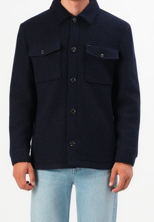 Mand iført en mørk navy uldjakke med knapper og klaplommer på brystet, kombineret med lyseblå jeans, stående foran en ensfarvet baggrund.