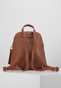 Zaino in pelle marrone con superficie testurizzata, forma arrotondata, doppie cinghie regolabili, manico superiore e dettagli in metallo color oro.