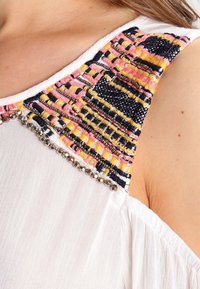Tessuto bianco con una yoke colorata e testurizzata che presenta fili rosa, gialli e blu navy, accentuata da perline metalliche lungo il collo.