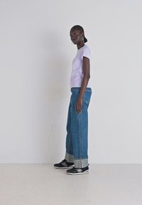 T-shirt fitted di un leggero viola, jeans blu a vita alta e larghi con orli arrotolati, e sneakers nere con strisce bianche. Tessuto liscio e texture denim.