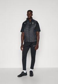 Schwarze, gesteppten Weste über einem schwarzen Kurzarmhemd, kombiniert mit enganliegenden schwarzen Hosen und schwarzen Sneakern, mit einem Nike-Logo und dem Emblem der Spurs.