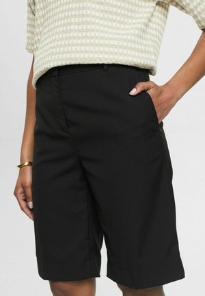 Black tailored shorts with a smooth texture, featuring side pockets and a clean hem. Paired with a light patterned top.
