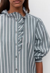 Camicia a righe grigio chiaro e bianca, con un particolare dettaglio a volant lungo il colletto e la lista dei bottoni, e maniche a sbuffo.