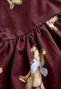 Tissu bordeaux avec des détails froncés mettant en avant des illustrations d'ours en peluche. Le tissu est doux, avec une texture lisse et des accents rayés bleus sur les ours.