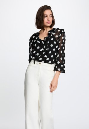 Femme portant un chemisier noir transparent à pois blancs et volants, associé à un pantalon large taille haute ivoire, debout devant un fond uni.