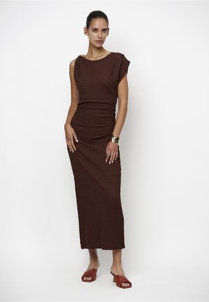 Woman wearing a sleeveless textured brown midi dress with a gold cuff bracelet and rust-colored heeled slide sandals, standing against a white background.
