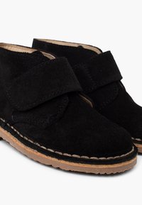 Bottines en daim noires avec une sangle Velcro, bout arrondi et détails decoutures autour de la semelle, dotées d'une semelle en caoutchouc brun clair.