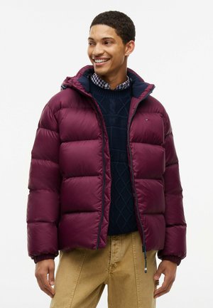 Mann lächelt, trägt eine dunkelrote Daunenjacke über einem marineblauen Pullover und beigen Hosen, schaut seitlich vor einem einfarbigen Hintergrund.