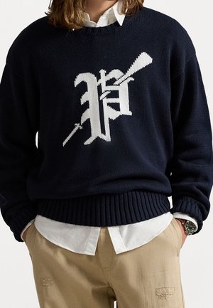 Persoon die een marineblauwe gebreide trui draagt met een witte gotische letter "P" en pijlenontwerp, wit overhemd, beige broek en polshorloge.