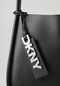 Svart teksturert lærhåndveske med lang stropp. Har en fremtredende sølv DKNY-logo-plakett på en matchende tag. Minimalistisk design.