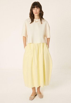 Kvinde med brunt hår iført en hvid top med korte ærmer, gul midi-nederdel, beige flettede flade sko og et guldhalskæde, står med hænderne i lommerne.