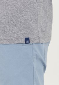 Tricou gri din bumbac cu etichetă albastră la margine, purtat cu pantaloni albaștri deschis. Materialele par moi și netede.