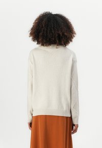 Person mit lockigem Haar, der einen cremefarbenen Strickpullover und einen orangen Rock trägt und mit dem Rücken zu einem schlichten weißen Hintergrund steht.