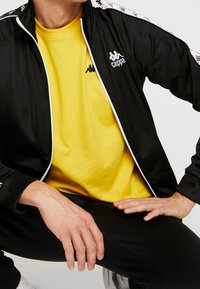 Muž v černé bundě Kappa přes žluté tričko, sedící s rukama opřenýma o stehna na jednobarevném pozadí.