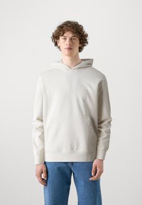 Lys beige hættetrøje med kangaroo-lomme og elastiske manchetter. Har et diskret logo på brystet, lavet af blødt stof. Sammen med blå jeans.