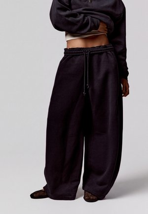 Personne portant un pantalon de jogging noir ample et un sweat-shirt noir court, assorti à des chaussures en mesh noires sur un fond uni.