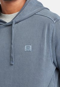 Sudadera con capucha en azul claro, de material texturizado. Cuenta con un parche de logo en el pecho, cordones en la capucha y un diseño casual.