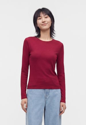 Jeune femme aux cheveux noirs mi-longs porte un t-shirt rouge ajusté à manches longues et un jean bleu clair, debout devant un fond blanc uni.