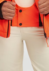 Veste rembourrée orange vif avec des boutons noirs, associée à un pantalon couleur crème. Des fermetures éclair et des cordons de serrage visibles soulignent le design.