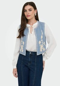 Ljust blå denimväst med vit blombroderi, scalloped kanter, bärs över en vit texturerad blus, tillsammans med mörkblå högmidjade jeans.