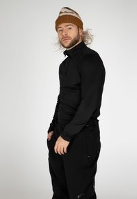 Maglietta nera a maniche lunghe con colletto a zip, abbinata a pantaloni neri. Un cappellino di maglia marrone con dettagli a motivo completa l'outfit.