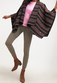 Brązowy i czarny w paski ponczo z szerokimi rękawami, noszone na różowej bluzce. Model łączy je z oliwkowymi wąskimi spodniami i brązowymi botkami za kostkę.